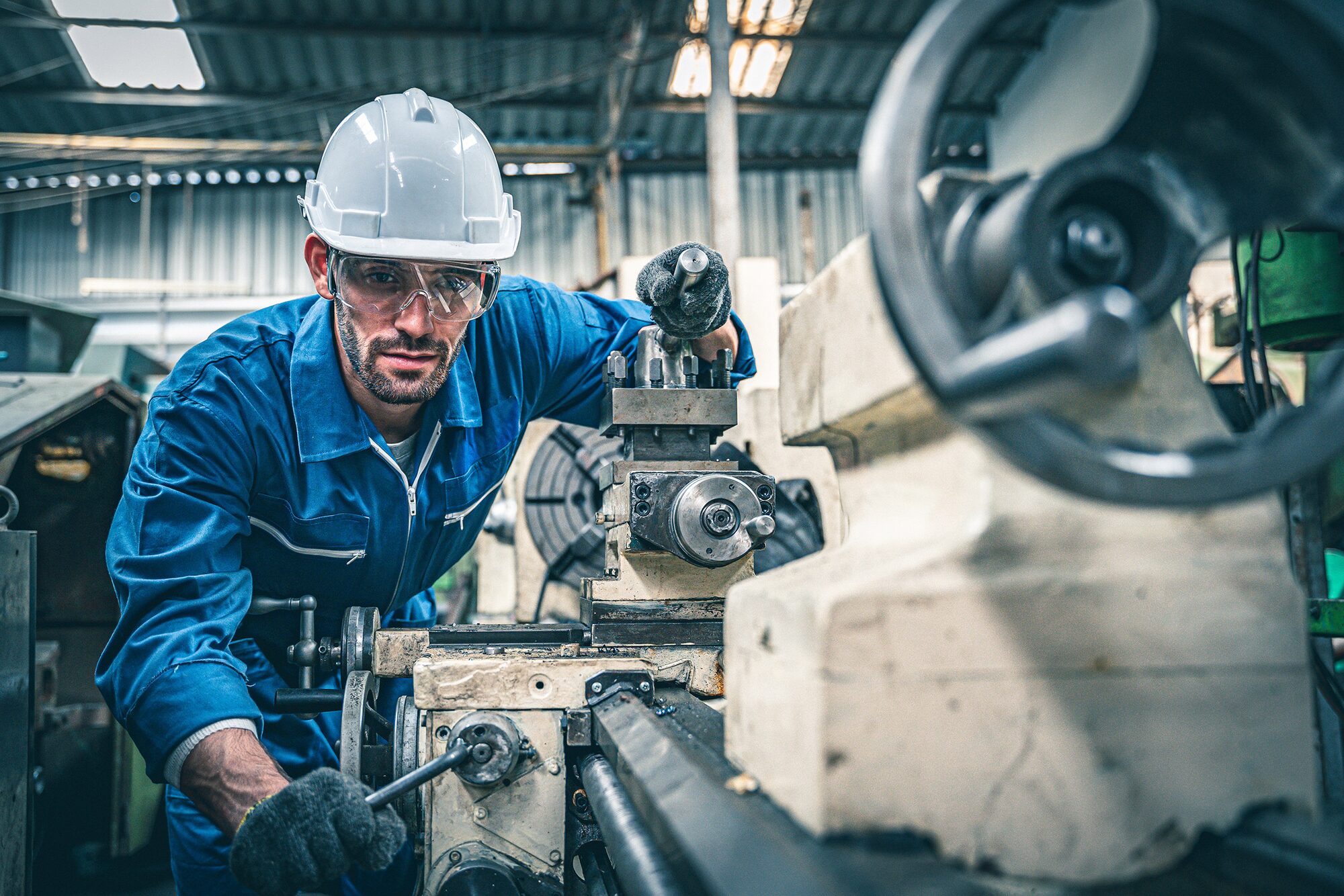 Worker in a blue jumpsuit and white hard hat operating a lathe